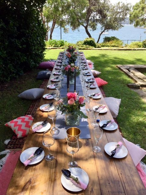 Boho Trestle Tables outside near the water with cushions on either side. Plates and flowers are top of the tables