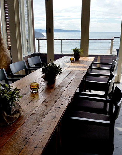 Vintage timber trestle table with black chairs on either side and a view of the ocean in the background