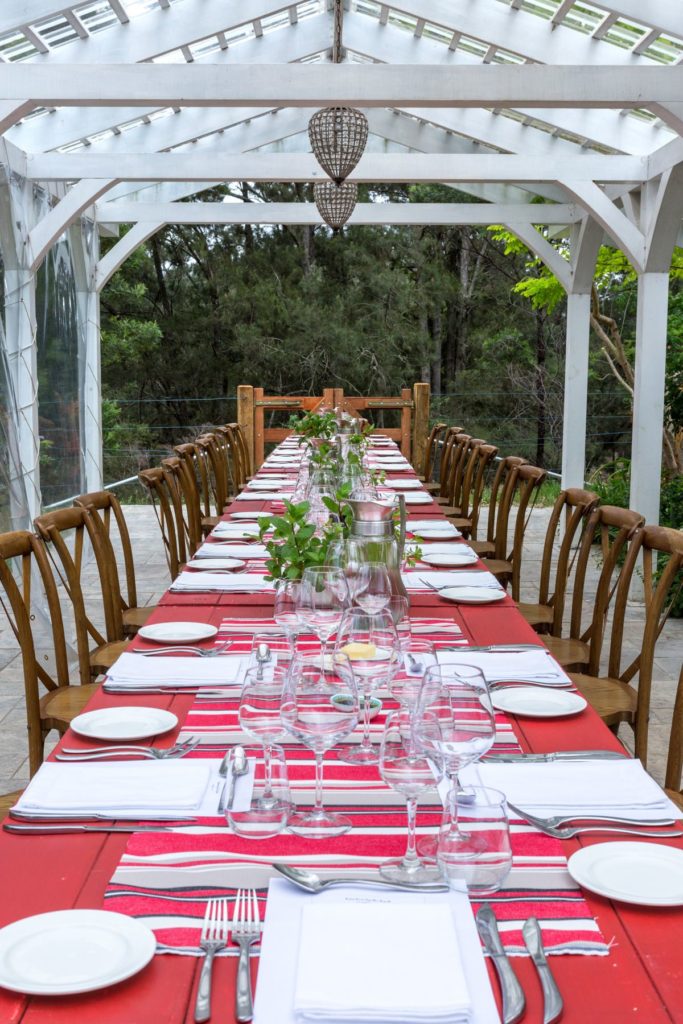Red painted trestle tables line dup with white plates, napkins, cutlery, wine glasses and small pnats down the centre of the tables. Wooden chairs are on either side, underneath a white roof outisde
